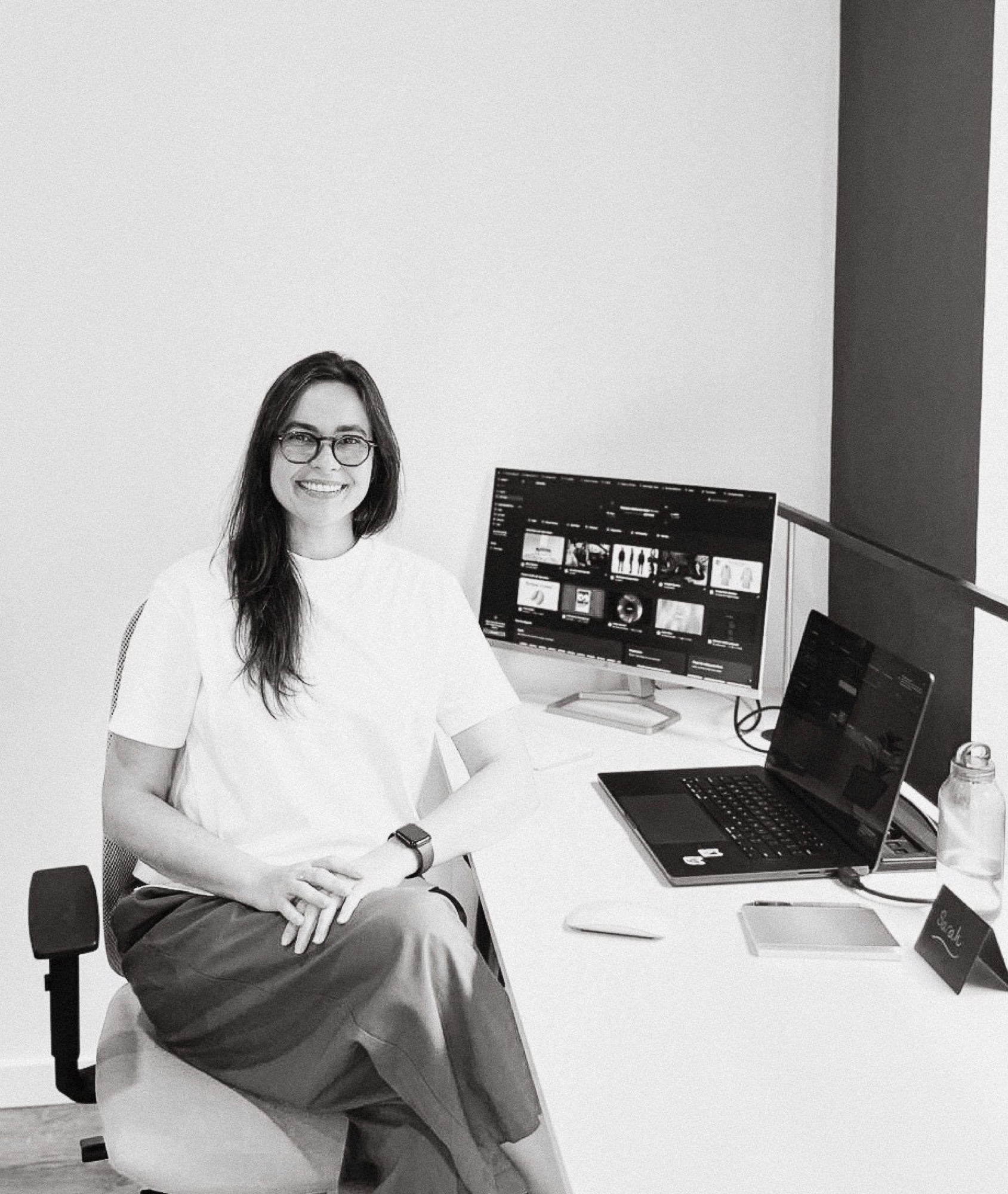 Sarah smiling at her desk, next to a monitor and a laptop